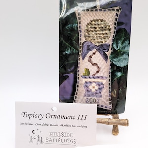 May include: A cross-stitch kit titled Topiary Ornament III by Hillside Samplings. The kit includes a chart, fabric, threads, a silk ribbon bow, and a small frog. The finished ornament features a topiary in a blue pot with the year 2003.
