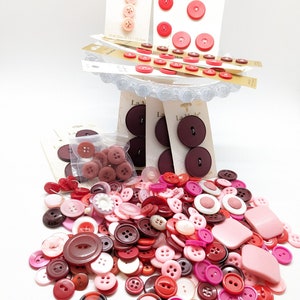 May include: A large collection of assorted buttons in various shades of pink, red, and brown. The buttons are arranged in a pile on a white surface. Some of the buttons are flat, while others are raised. There are also some buttons with a decorative design.