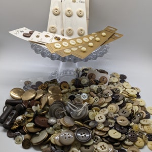 May include: A large collection of assorted buttons in various shades of brown, beige, and black. The buttons are arranged in a pile on a white surface. There are also several packages of buttons in the background, including a package labeled "Belle Buttons by Dritz".