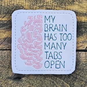 Könnte beinhalten: Weißer gestickter Aufnäher mit einem rosa Gehirngraphic und blauem Text, der "Mein Gehirn hat zu viele Tabs offen" lautet.