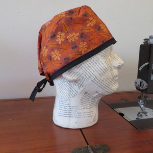 Puede incluir: Un gorro de quirófano con un estampado de otoño de hojas naranjas, amarillas y marrones sobre un fondo color óxido. El gorro tiene un ribete negro y lazos negros.