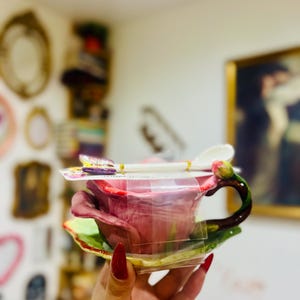 May include: A pink rose-shaped teacup and saucer set with a small spoon. The set is wrapped in plastic and has a green leaf design on the saucer.