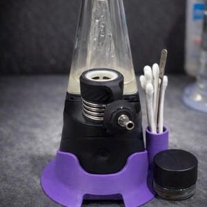 May include: A black and purple electronic device with a clear glass component. Accessories include a small glass jar with a black lid, a purple holder with cotton swabs, and a metal tool. The device has a modern design.