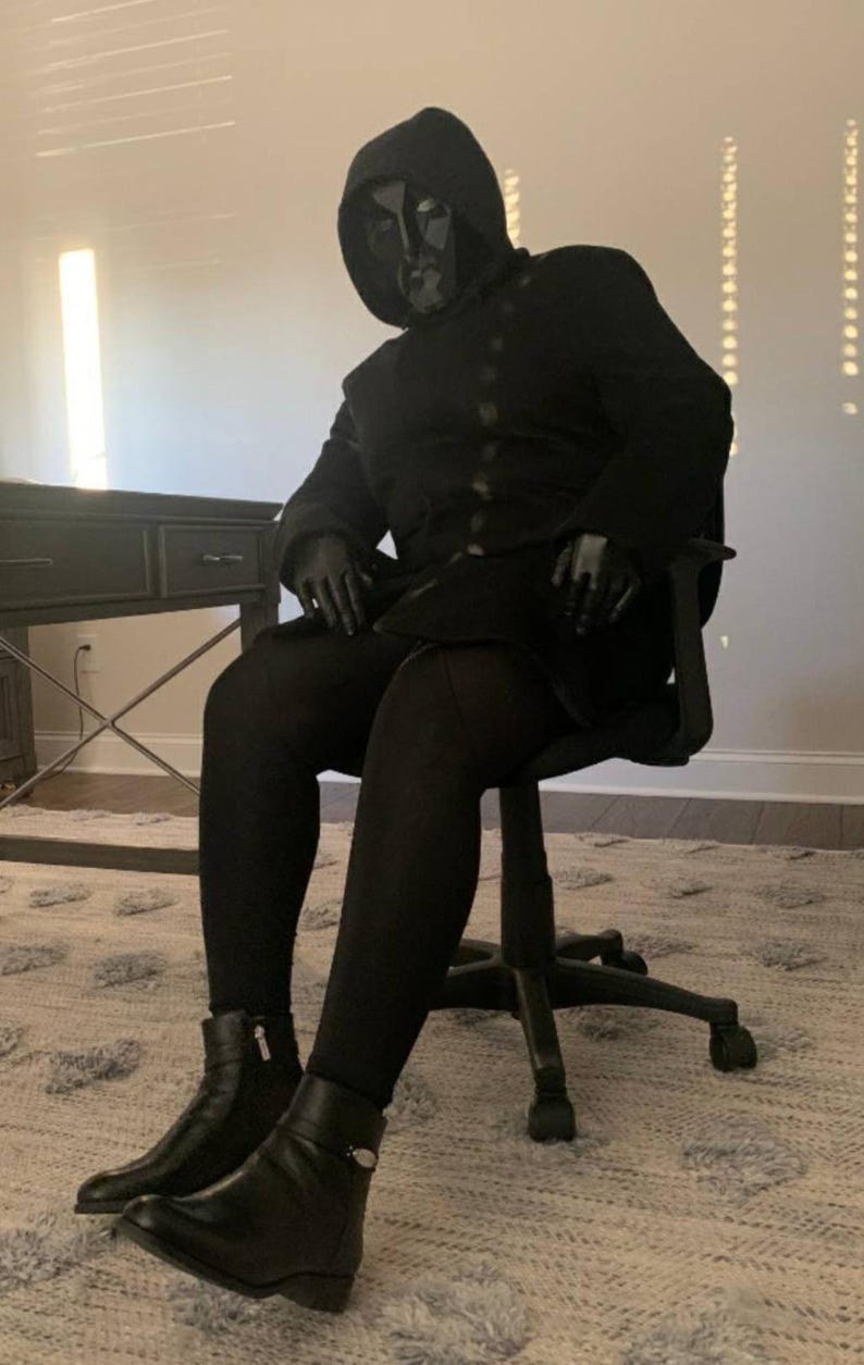 K&ouml;nnte beinhalten: Eine Person in einem schwarzen Kapuzenmantel und einer Maske sitzt in einem schwarzen B&uuml;rostuhl. Die Person tr&auml;gt schwarze Handschuhe, eine schwarze Hose und schwarze Lederstiefel. Der Hintergrund umfasst einen Schreibtisch und einen gemusterten Teppich.