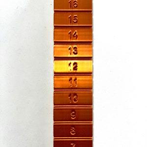 May include: A copper-colored metal ruler with numbers 6 through 18 marked on each section. The ruler is a vertical strip with each number on a separate section.