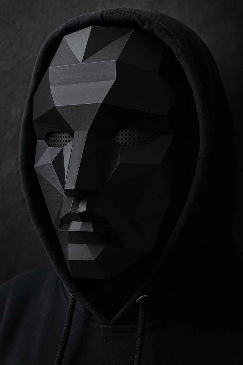 K&ouml;nnte beinhalten: Eine schwarze geometrische Maske und ein schwarzer Kapuzenpullover. Die Maske hat ein polygonales Design mit Augen- und Mund&ouml;ffnungen. Die Maske ist dunkel und der Pullover ist schwarz.