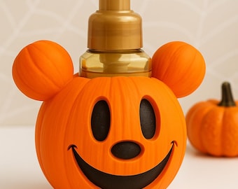 May include: Orange Halloween-themed soap dispenser shaped like a pumpkin with a smiling face and Mickey Mouse ears. The dispenser has a gold-colored pump and a small pumpkin in the background. Perfect for Halloween decor.