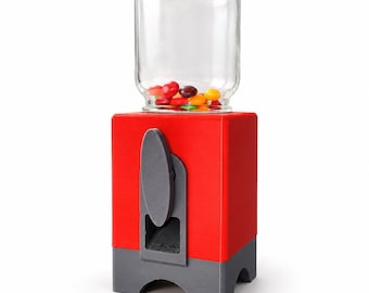 May include: A red and grey candy dispenser featuring a clear glass jar top filled with colourful sweets. The base is a rectangular red block with a grey bottom section and a grey manual dispensing lever on the front face for releasing treats.