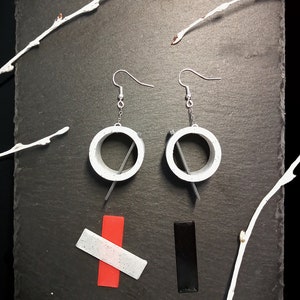 Può includere: Orecchini geometrici con cerchi bianchi e un accento grigio, con ganci argentati. Un rettangolo rosso e bianco e un rettangolo nero sono anch'essi presenti. Rami dipinti di bianco sullo sfondo.