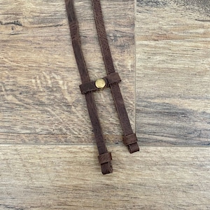 May include: Close-up of brown leather braces with a brass-coloured button clasp. The braces are made of distressed leather and are designed to be worn over the shoulders. The background is a wooden surface.