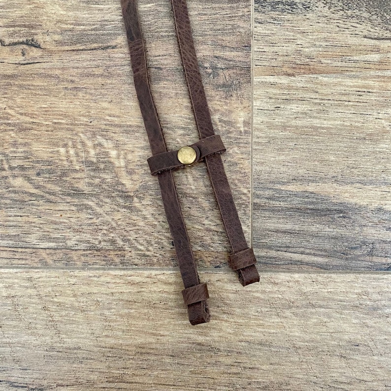 May include: Close-up of brown leather braces with a brass-coloured button clasp. The braces are made of distressed leather and are designed to be worn over the shoulders. The background is a wooden surface.