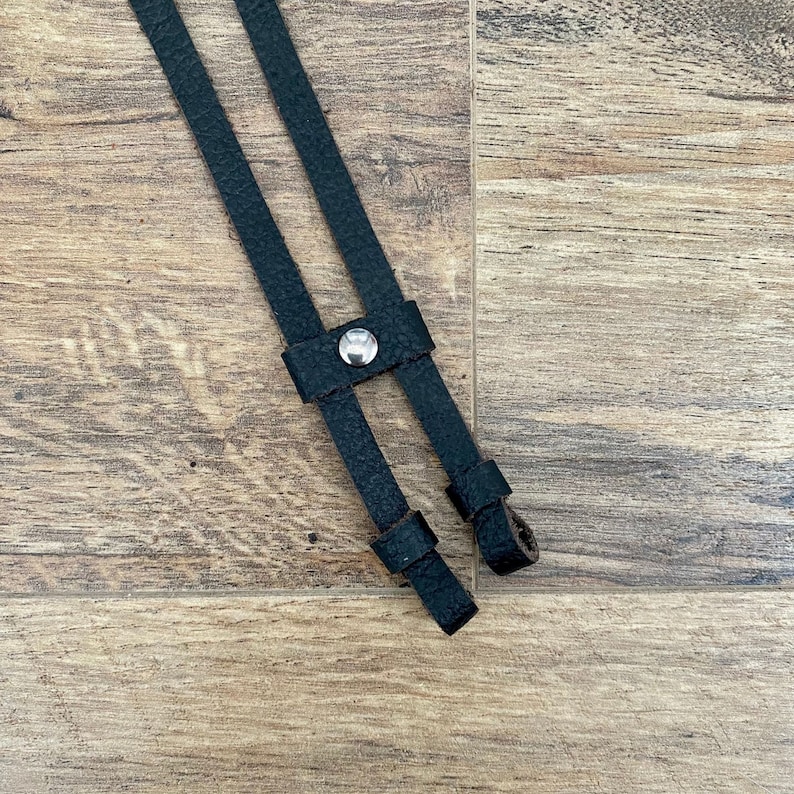 May include: Close-up of black leather suspender straps with a silver button detail. The straps are textured and appear to be attached. The background is a light brown wooden surface.