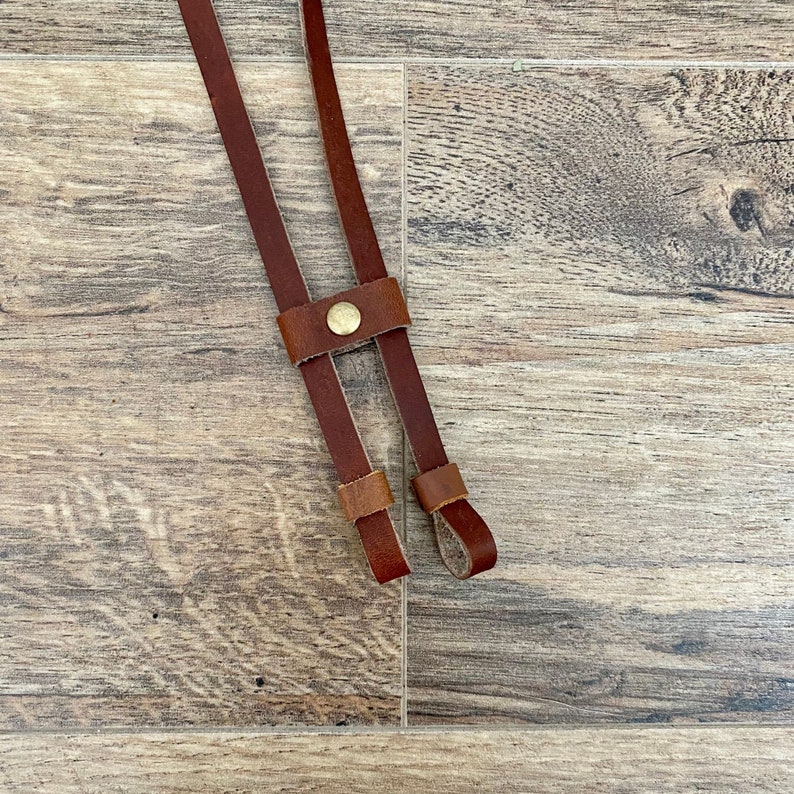 May include: Close-up of a brown leather strap with a brass button and two smaller loops. The strap is designed with two parallel sections connected by a buttoned piece. The background is a wooden surface.