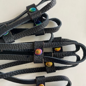 May include: Close-up of several black leather straps with iridescent metal rivets. The straps are arranged in a layered pattern, with the rivets adding a pop of colour. The leather has a textured appearance.