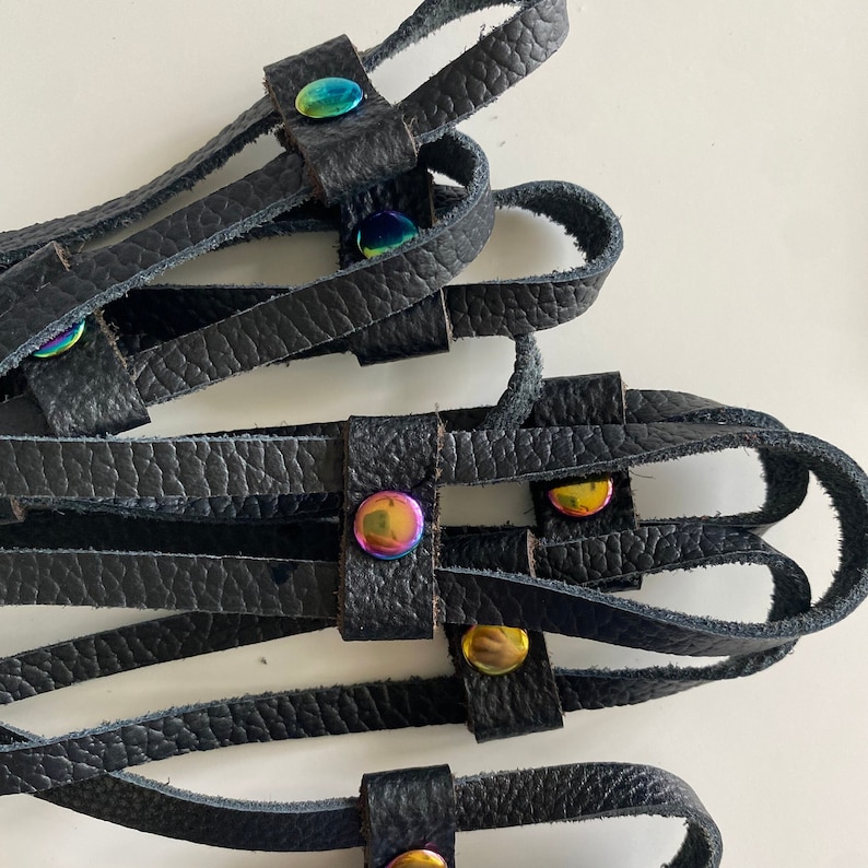 May include: Close-up of several black leather straps with iridescent metal rivets. The straps are arranged in a layered pattern, with the rivets adding a pop of colour. The leather has a textured appearance.