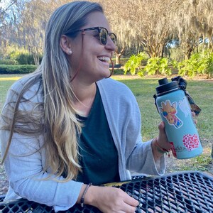 May include: A light blue water bottle adorned with stickers, including a giraffe in sunglasses and a red heart design. The bottle has a black lid and strap. The person is wearing sunglasses and a grey cardigan. The background shows trees.