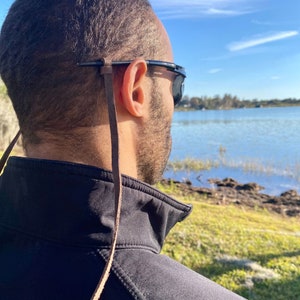 May include: A person wearing black sunglasses with a brown leather eyeglass strap. The strap is attached to the temples of the glasses and hangs down. The background shows a lake and blue sky.