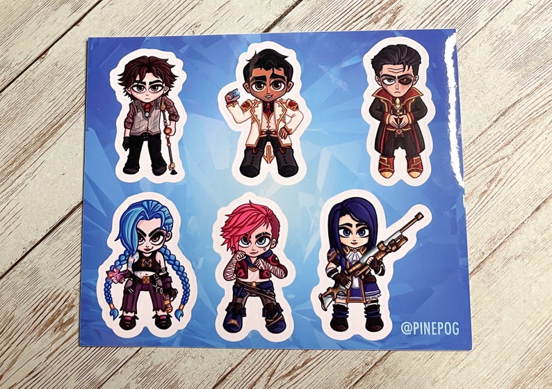 May include: A set of six colorful cartoon character stickers featuring characters from the video game League of Legends. The stickers are printed on white paper with a blue background. The characters include Jinx, Vi, Jayce, Caitlyn, Viktor, and Ekko. The text "@PINEPOG" is printed in the bottom right corner.