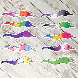May include: A selection of worm stickers, each displaying a different pride flag design. The stickers feature cartoon worms with smiling faces, in a variety of colours, including rainbow, lesbian, transgender, and non-binary flags.
