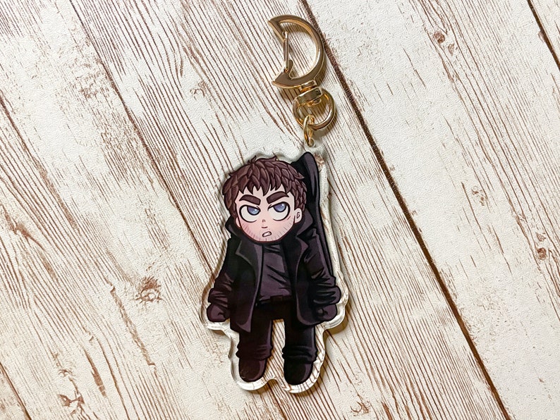 Puede incluir: Un llavero con un personaje de dibujos animados en blanco y negro con un llavero de metal dorado. El personaje es un hombre con cabello casta&ntilde;o y una chaqueta negra. Lleva pantalones negros y zapatos negros.