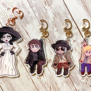 Puede incluir: Cuatro llaveros de personajes de dibujos animados coloridos con cierres de metal dorado. Los personajes son una mujer con un vestido blanco y un sombrero negro, un hombre con un abrigo negro, un hombre con un abrigo marr&oacute;n y gafas de sol, y un hombre con una chaqueta blanca y pantalones vaqueros azules.
