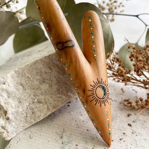 May include: A handcrafted wooden tool with a Y-shape, featuring a sun and moon design. The light brown wood is adorned with turquoise and white dot accents. The tool is likely used for massage or self-care.