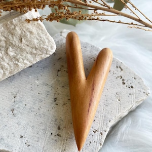 Könnte beinhalten: Ein hellbraunes Gua-Sha-Werkzeug aus Holz in V-Form, das auf einem strukturierten grauen Stein liegt. Das Werkzeug hat eine glatte, polierte Oberfläche. Getrocknete Pflanzen und eine weiße, flauschige Oberfläche befinden sich im Hintergrund.
