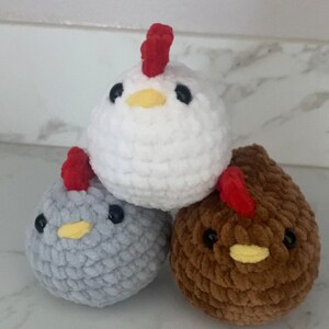 May include: Three crocheted plush chickens, one white, one gray, and one brown. Each chicken has a red comb and black eyes.