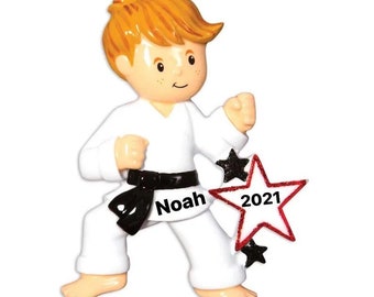 May include: White personalized Christmas ornament of a boy in a karate uniform with a black belt. The ornament has a red star with black glitter and the year 2021. The boy's name, Noah, is written on the belt.