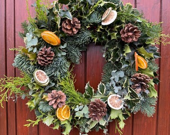 May include: A festive Christmas wreath with a mix of greenery, including holly, pine, and eucalyptus. The wreath is adorned with pine cones and dried orange slices, creating a natural and seasonal look. A green ribbon is used for hanging.