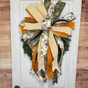 May include: A decorative wreath featuring a large, multi-layered bow made of various ribbons. The ribbons include patterns of yellow and white geometric circles, green and brown plaid, and a floral print with autumn-colored leaves on a white background.