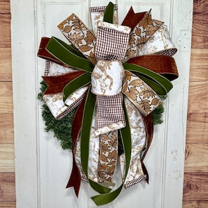 May include: A decorative wreath bow featuring a mix of ribbons in brown and white gingham, solid olive green velvet, and a white fabric with a brown autumn leaf pattern. The bow has multiple loops and long, flowing tails arranged in a layered design.