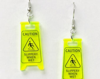 Pendientes extraños de acrílico con la leyenda "Precaución: resbaladizo cuando está mojado", señal de suelo mojado, regalo kitsch de seguridad.