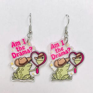 May include: A pair of novelty earrings featuring a cartoon frog wearing a cowboy hat and holding a heart-shaped mirror. The earrings have the text "Am I the Drama?" in pink. The frog is light green with dark green spots.