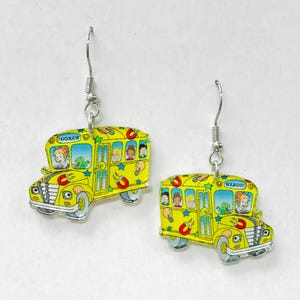 May include: A pair of yellow school bus earrings with silver-colored hooks. Each earring features a cartoon bus design with passengers and the words "OOOHAW" and "WAHOO!" in the top corners. The earrings have a glossy finish.