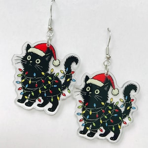 Op de afbeelding: Een paar kerstoorbellen met illustraties van zwarte katten. Elke kat draagt een rode kerstmuts en is gewikkeld in kleurrijke kerstverlichting. De oorbellen hebben zilverkleurige oorhaken.