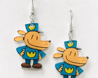 Op de afbeelding: Een paar oorbelhangers met een cartoonhond die een blauw uniform draagt met een gele kroon. De hond heeft een bruin lichaam, een wit gezicht en een zwarte neus.