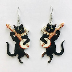 Puede incluir: Un par de pendientes originales con figuras de gatos negros tocando el banjo. Los gatos llevan cuellos con volantes y tienen ojos amarillos. El banjo tiene una cara blanca y un mástil rojo y marrón. Los pendientes cuelgan de ganchos plateados.