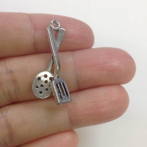 May include: Silver charm featuring a crossed spatula and slotted spoon. The charm is designed to look like kitchen utensils.