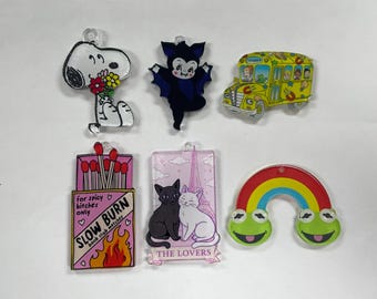 May include: A collection of colorful acrylic charms featuring various designs. Includes a Snoopy-like character holding flowers, a vampire, a yellow school bus, a matchbox with text, a cat-themed design, and a rainbow with frog faces.
