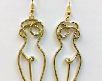 May include: A pair of gold-colored wireframe earrings. Each earring is shaped like a stylized female figure, with a looped head and a curved body. The earrings hang from gold-colored fish hook ear wires.