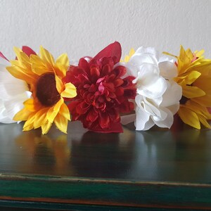 Puede incluir: Una corona floral con girasoles, flores blancas y flores rojas. Los girasoles tienen pétalos amarillos y centros marrones. Las flores blancas están agrupadas y las flores rojas son de color carmesí intenso. La corona es para una ocasión festiva.