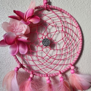 Pode incluir: Apanhador de sonhos rosa com penas rosa e um enfeite de flores rosa. O apanhador de sonhos tem um amuleto prateado com a palavra "Libra" nele.