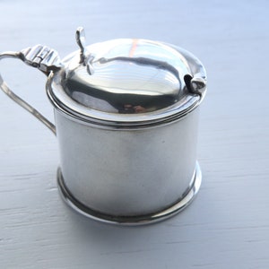 May include: A small, silver-coloured container with a lid and handle. The container has a cylindrical body and a rounded lid with a small handle. The handle is curved and attached to the side. The lid has a small opening.