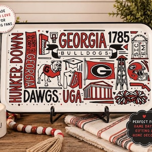 May include: A rectangular white serving tray with black and red graphic illustrations featuring the Georgia Bulldogs logo, a bulldog mascot, the text Georgia 1785, Hunker Down, and Sic Em. The tray has a black rim and two wooden handle grips.