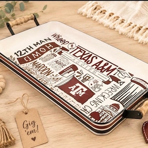 May include: A rectangular white serving tray with maroon and black graphics featuring Texas A&M university themes, including the text 12TH MAN, WHOOP, AGGIES, and WRECKING CREW. The tray has black metal handles with light wood accents on each side.