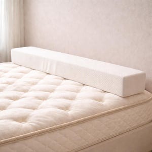 May include: A long, rectangular white foam bed bumper sits atop a quilted mattress. The bumper is made of a textured material and is designed to prevent a person from rolling off the bed. The mattress is a light cream color.