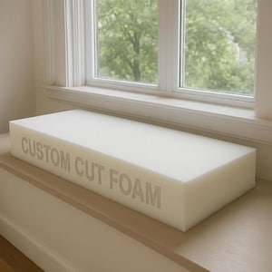 May include: A rectangular block of white foam with the text "CUSTOM CUT FOAM" printed on the side. The foam is placed on a wooden surface near a window with a view of green trees.