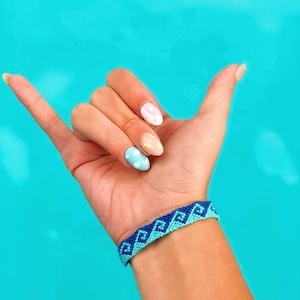 May include: A hand with blue, white, and yellow nail polish with a blue and white friendship bracelet on the wrist.