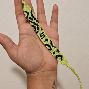 Peut inclure: Un bracelet d'amitié fait à la main, vert citron avec des lettres noires épelant un mot. Le bracelet est tenu dans une main sur un fond neutre. Le bracelet est fait de fils tissés.